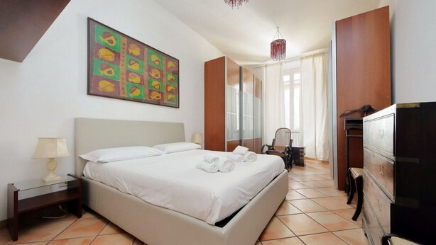 Een hotelkamer met een bed, nachtkastje, stoel, kapstok en een schilderij aan de muur.