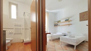 Een badkamer met douche, toilet en wastafel. Er staat een bed met witte lakens en een nachtkastje.