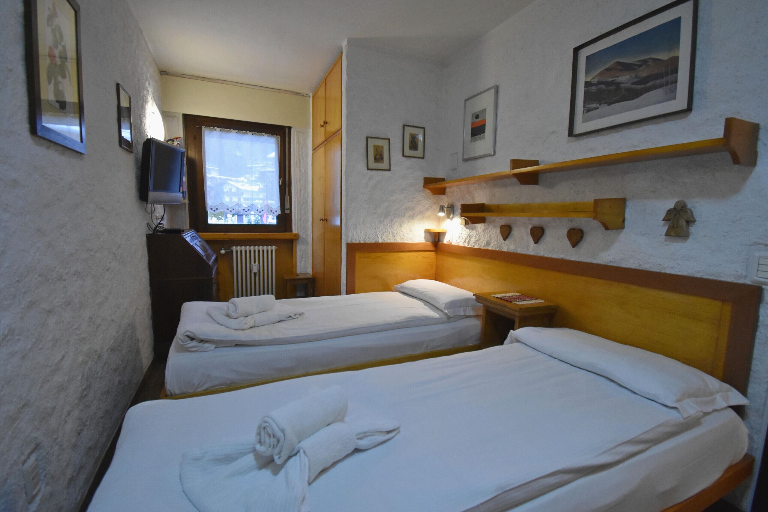 A hotel room with two beds, a TV, a window, and a wooden shelf with decorative items.