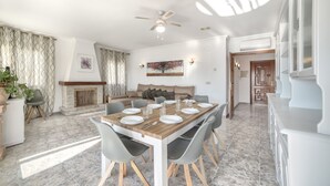 A dining area with a wooden table set for six, surrounded by grey chairs. A fireplace is visible in the background, and the room has a ceiling fan. - Villa Garrofer- PlusHolidays (Calpe)