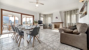 A spacious living area with a dining table, chairs, a sofa, a fireplace, and large windows. - Villa Garrofer- PlusHolidays (Calpe)