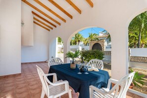 A covered outdoor dining area with white chairs and a table set for two, featuring a view of palm trees and a stone building in the background.