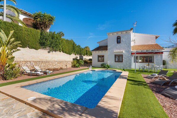 A swimming pool with lounge chairs and a white house with a tiled roof.