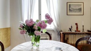 Ein Esstisch mit Blumen in einer Vase, ein gerahmtes Bild an der Wand und ein Fenster mit Vorhängen.