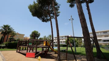 Children can play on the playground equipment, including a play structure with slides and swings, surrounded by lush grass and trees.