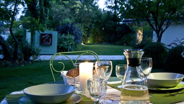 An outdoor dining setup with a candle, wine glasses, and a bottle, surrounded by greenery and a sign in the background. - YOUR HOME AWAY FROM HOME (Treviso)