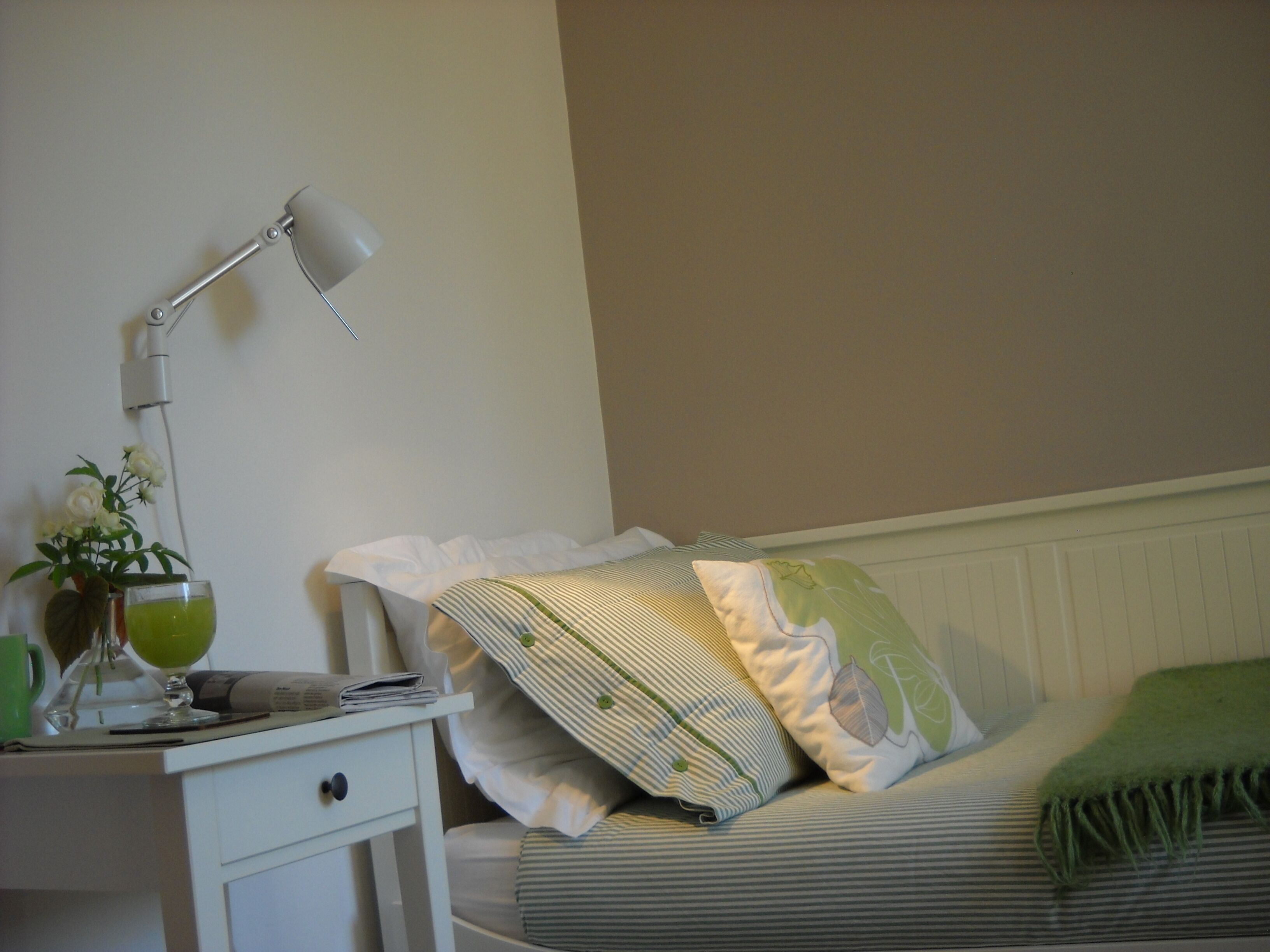 Een opgemaakt bed met gestreepte lakens en een groene plaid, een nachtkastje met een lamp, een vaas met bloemen en een kop.