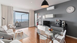Uma sala de estar moderna com mesa de jantar de vidro, cadeiras brancas, um sofá e uma TV.