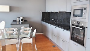 Uma cozinha moderna com mesa de jantar de vidro, cadeiras brancas, eletrodomésticos de aço inoxidável e armários cinzentos.