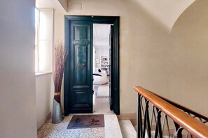 Un pasillo con una puerta de madera oscura, un cubo metálico, un felpudo y una escalera con barandilla de madera.