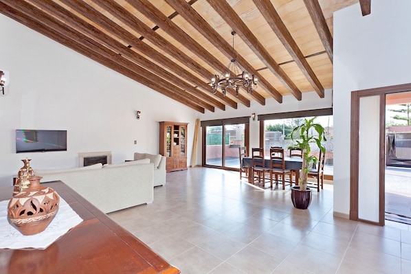 A spacious living area with a wooden ceiling, a white sofa, a dining table with chairs, and a potted plant.