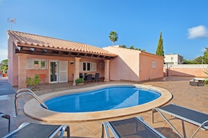 A swimming pool with lounge chairs, a patio with a table and chairs, and a building with a tiled roof.