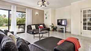 Ett modernt vardagsrum med en soffa, ett soffbord och en tv. Det finns en balkong med gardiner och utsikt över grönområden.