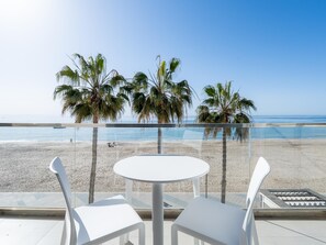 Un balcón con una mesa blanca y dos sillas blancas, con vista a una playa con palmeras y el océano.
