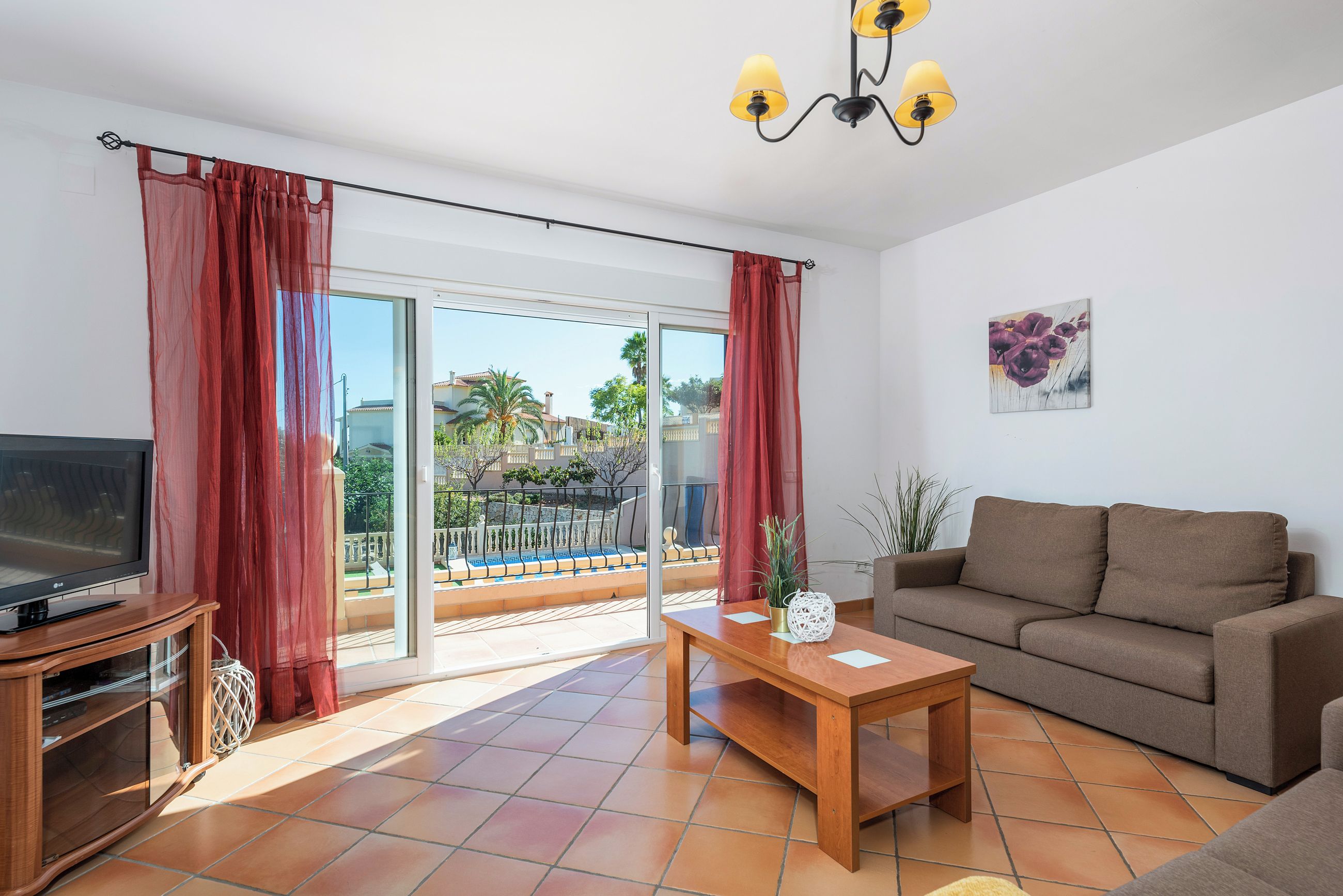 Een woonkamer met een bank, een televisie, een houten salontafel en een balkon met rode gordijnen.