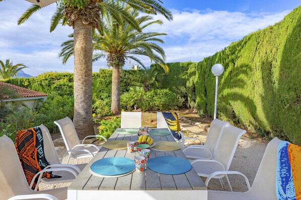 An outdoor dining area with a table set for four, surrounded by white chairs, palm trees, and a lush green hedge.