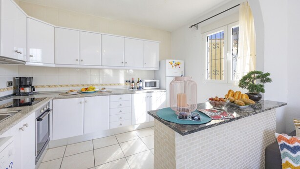 A modern kitchen with white cabinets, a built-in oven, a microwave, and a breakfast bar with pastries and a coffee maker. - Villa Carola - PlusHolidays (Calpe)