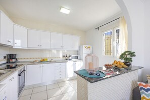 Una cocina moderna con armarios blancos, horno integrado, microondas y una barra de desayuno con pasteles y una cafetera.