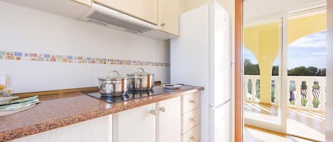 A kitchen with white cabinets, a granite countertop, and a white refrigerator. There are pots on the stove and a window overlooking a balcony.