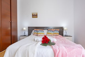 Ein ordentlich bezogenes Bett mit weißen Leinen, zwei Kissen mit Zickzackmuster, einer rosa Decke und einer roten Blumenkomposition.
