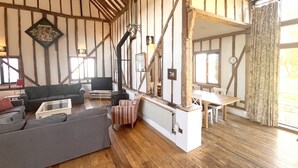 A spacious living area with a high ceiling, exposed wooden beams, a fireplace, and a dining area with a table and chairs. - The Dairy Hall at Partridge Lodge (Woodbridge)