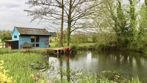 A small blue cabin by a pond with a wooden bench and a red ring on the wall. - The Dairy Hall at Partridge Lodge (Woodbridge)