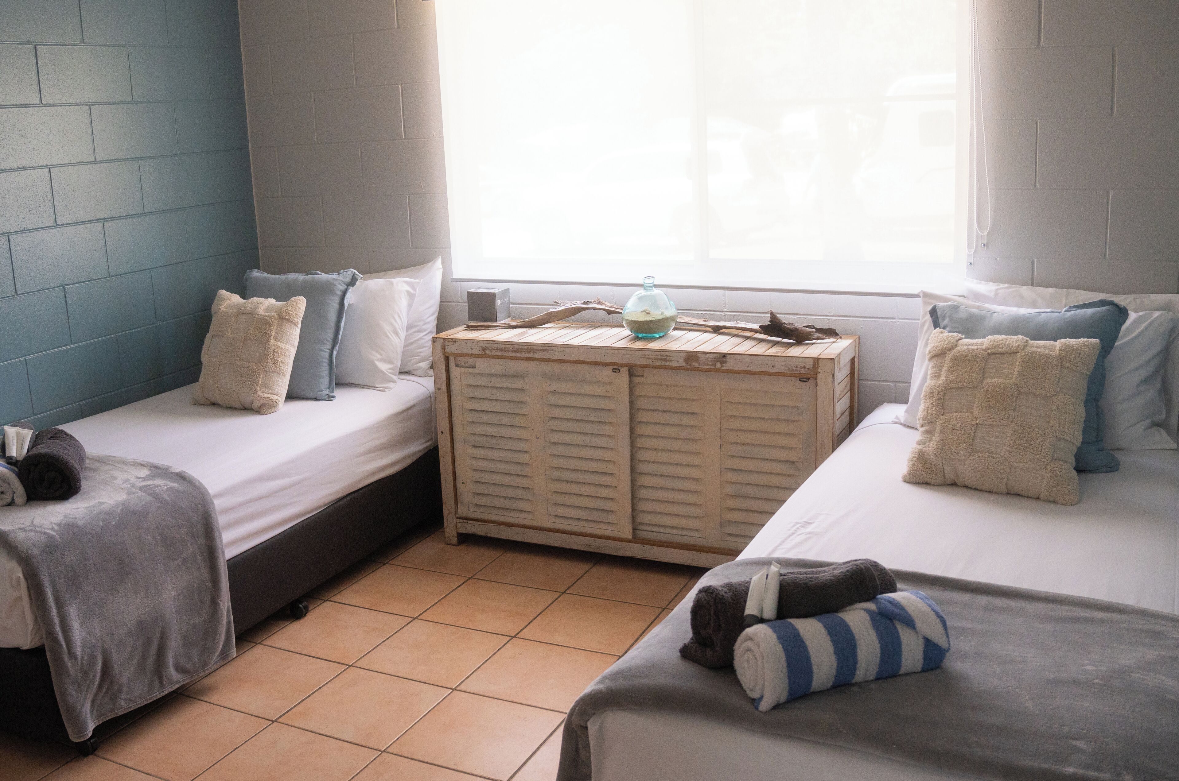 Een kleine, gezellige kamer met twee eenpersoonsbedden, een houten dressoir en een raam met gordijnen.