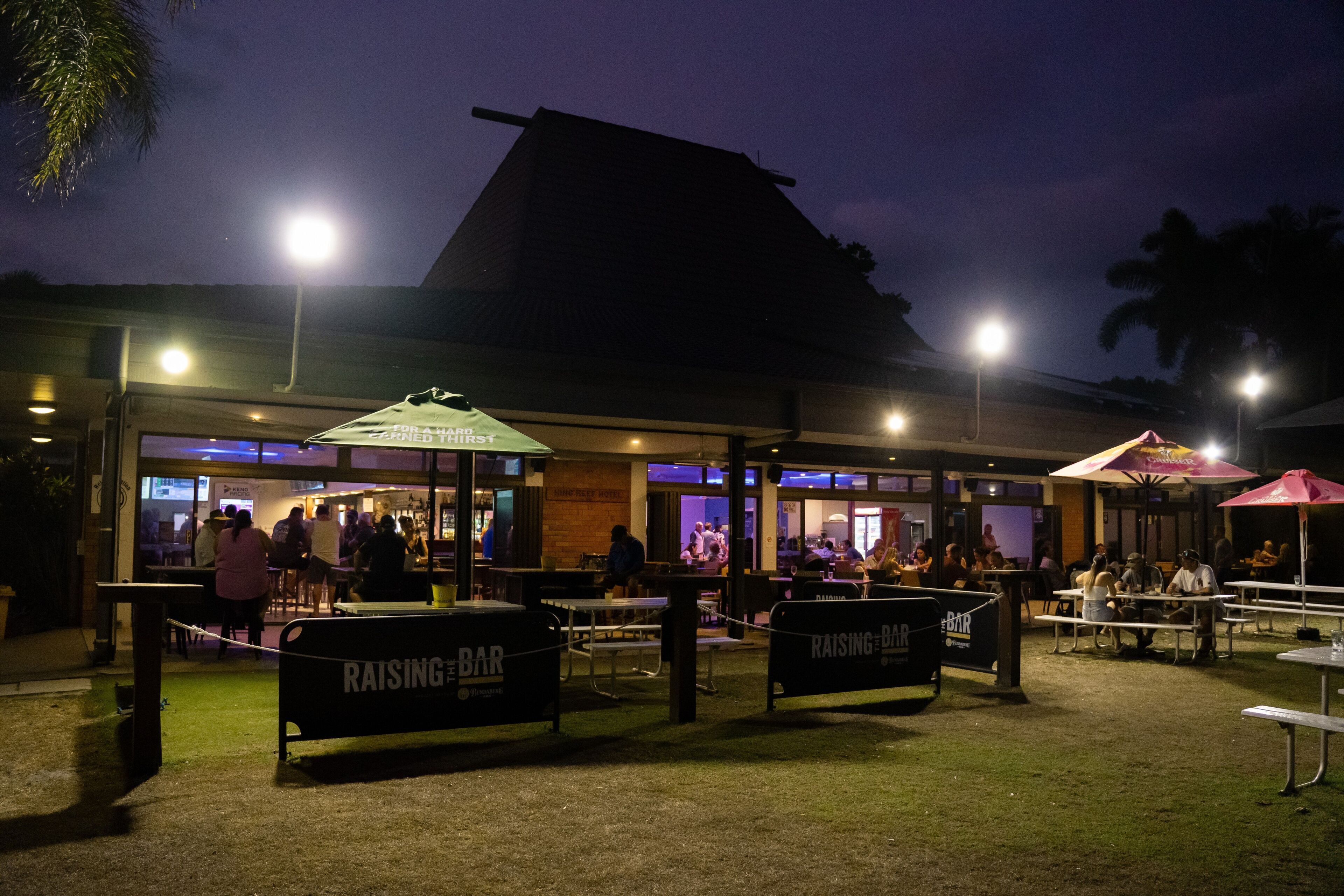 An outdoor dining area with tables and chairs, surrounded by a low fence, and illuminated by overhead lights.
