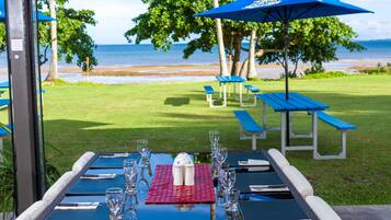 Un espacio de comedor al aire libre con mesas dispuestas para dos, sombrillas azules y vista a la playa.