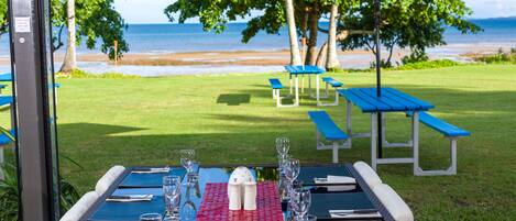 Un espacio de comedor al aire libre con mesas dispuestas para dos, sombrillas azules y vista a la playa.