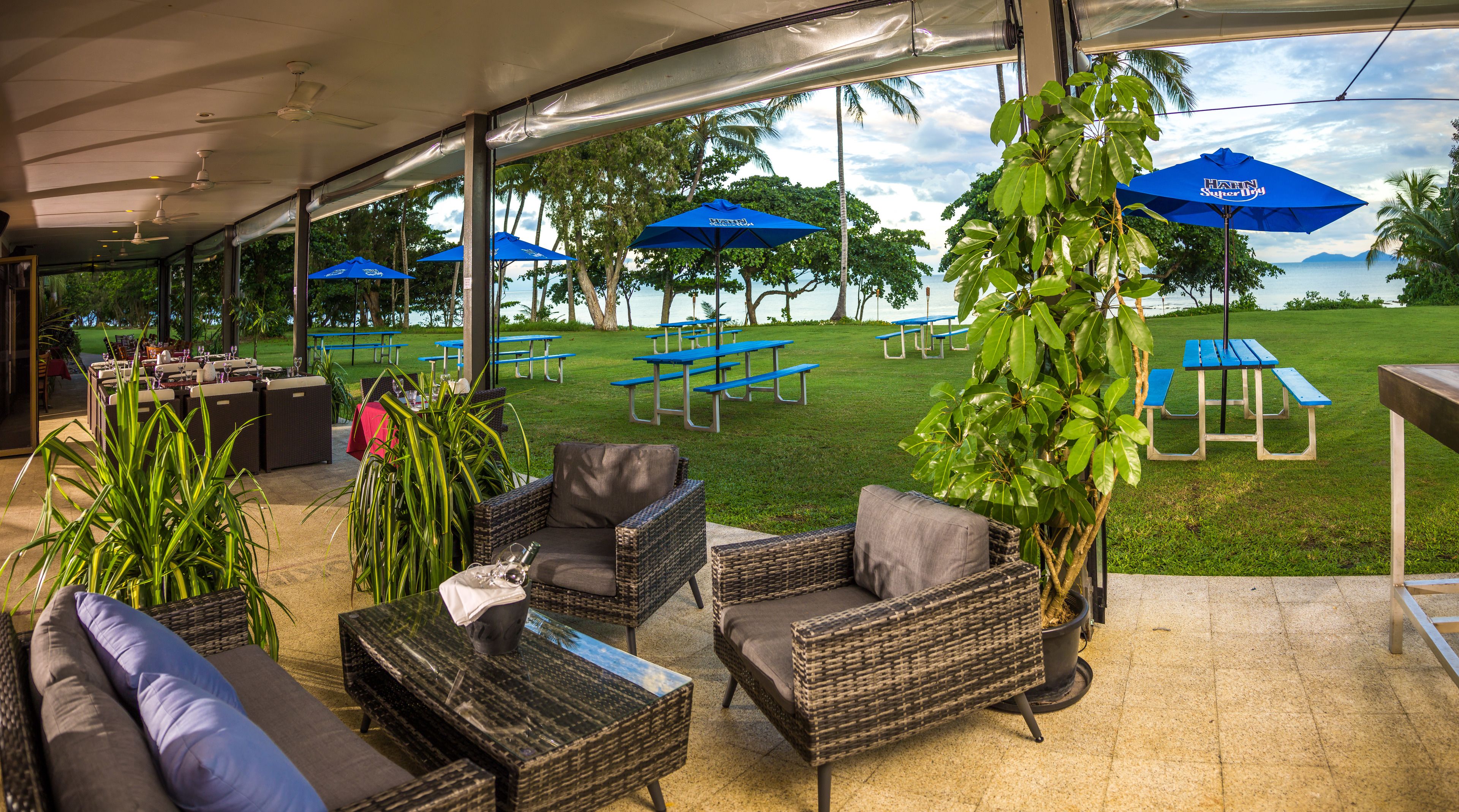 Eine Terrasse im Freien mit Korbmöbeln, blauen Sonnenschirmen und Picknicktischen.