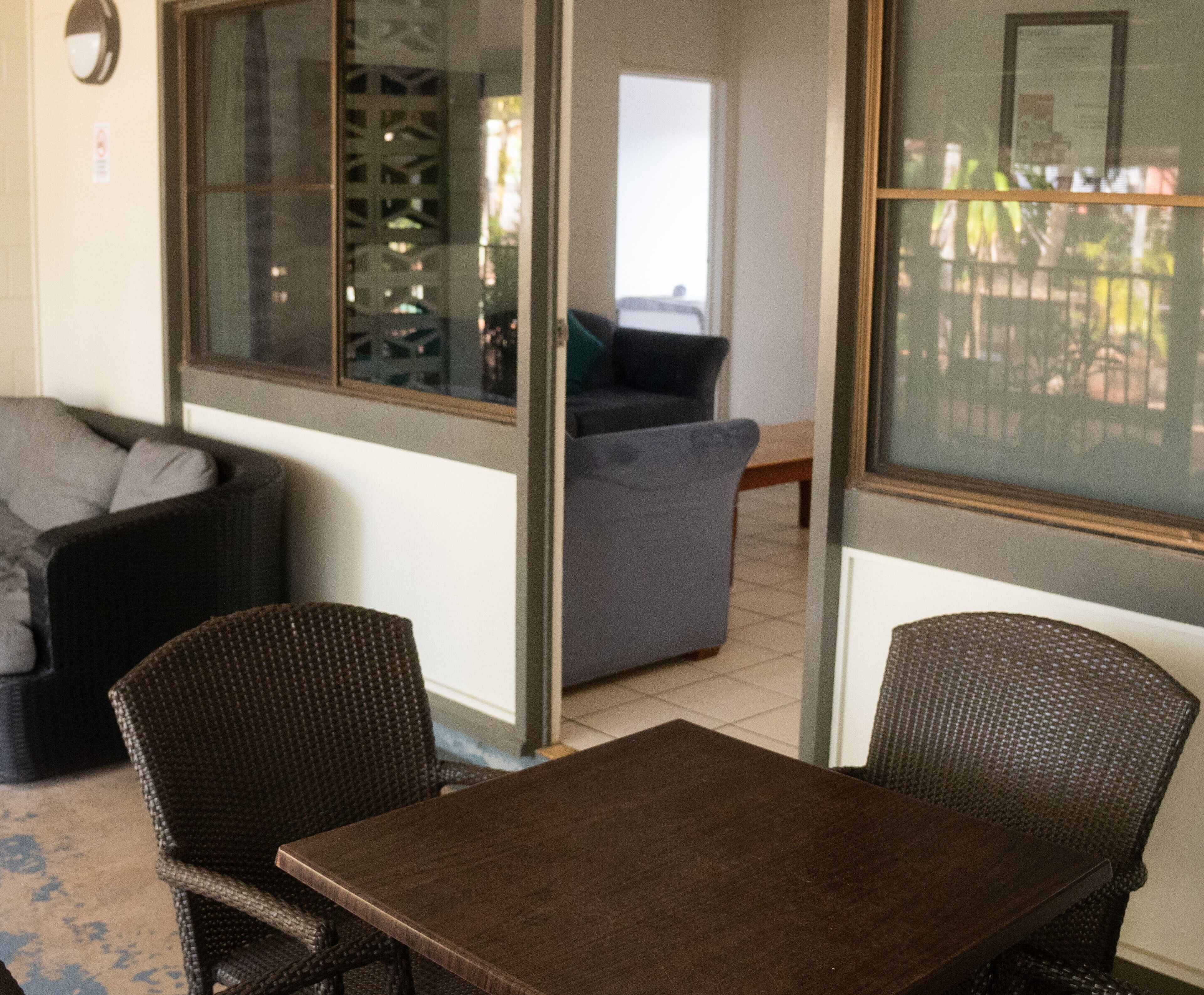 A room with a dark wooden table and two wicker chairs, a grey sofa, and a window with a view of a fence and greenery.