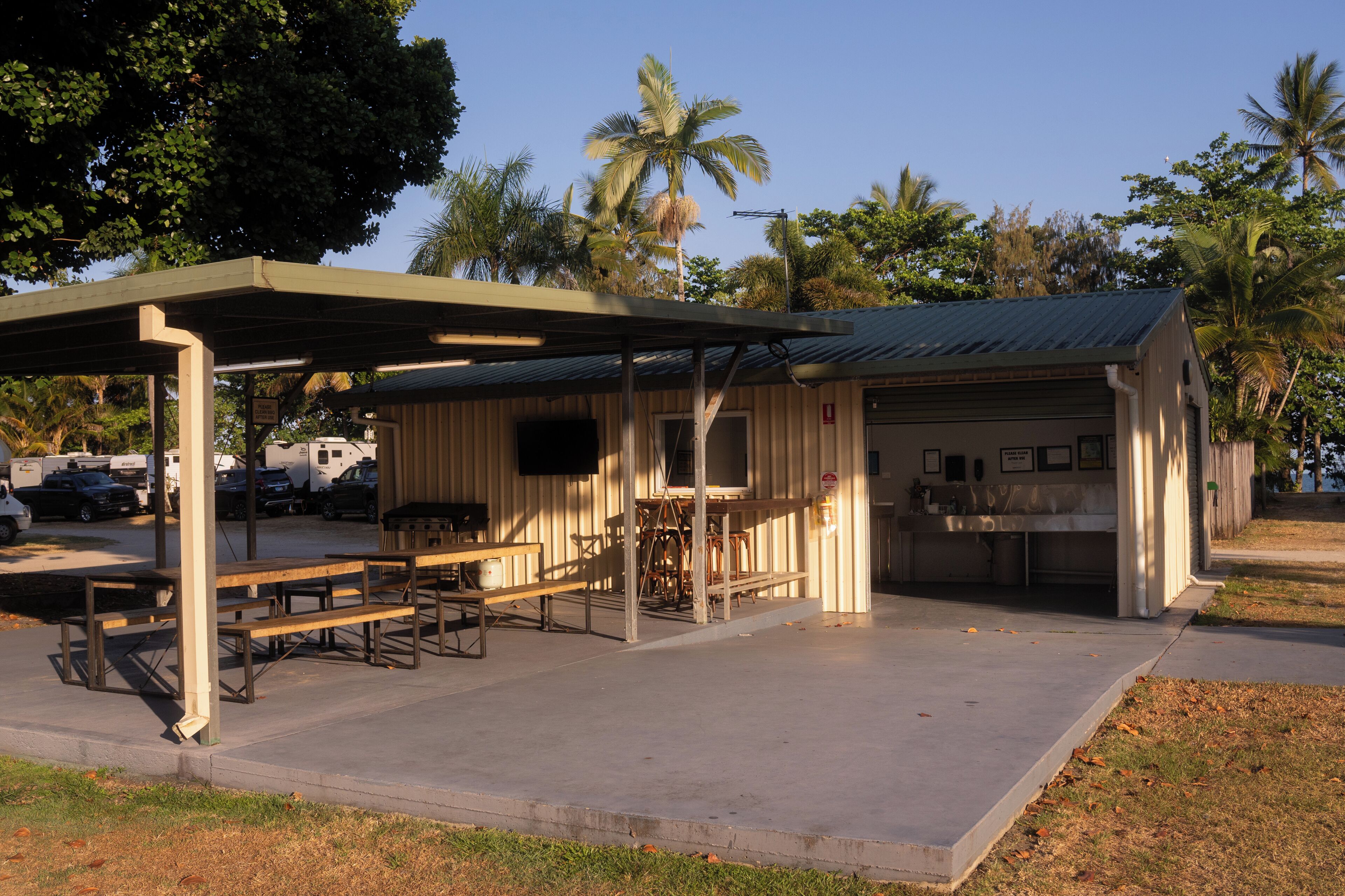 Un área de picnic techada con mesas y bancos, un pequeño edificio con cocina, y palmeras al fondo.