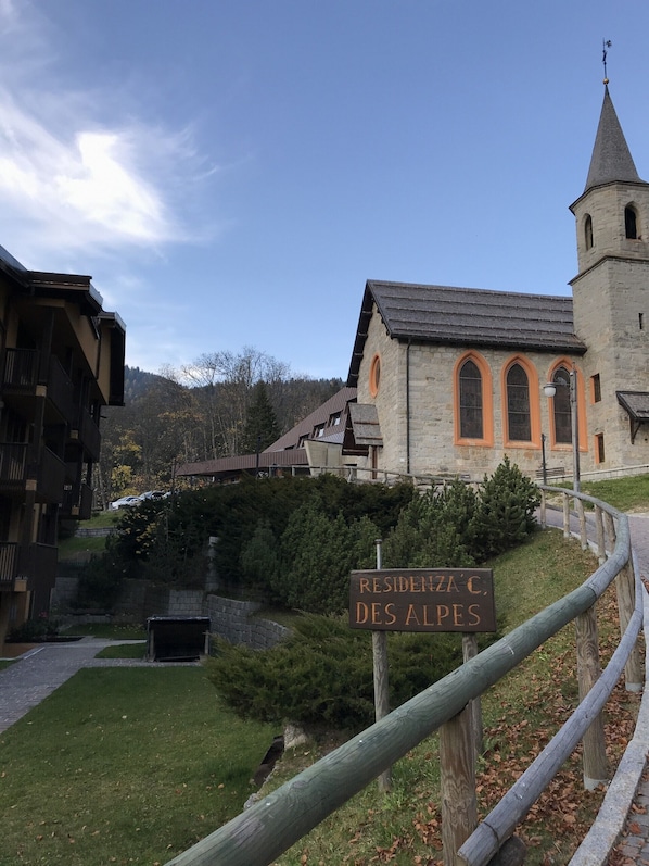 A sign reading "RESIDENZA C. DES ALPES" in front of a stone building with a tower, surrounded by greenery and a wooden fence.