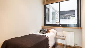 Une petite chambre à coucher, bien rangée, avec un lit simple, une table de chevet, une lampe et une fenêtre avec des stores.
