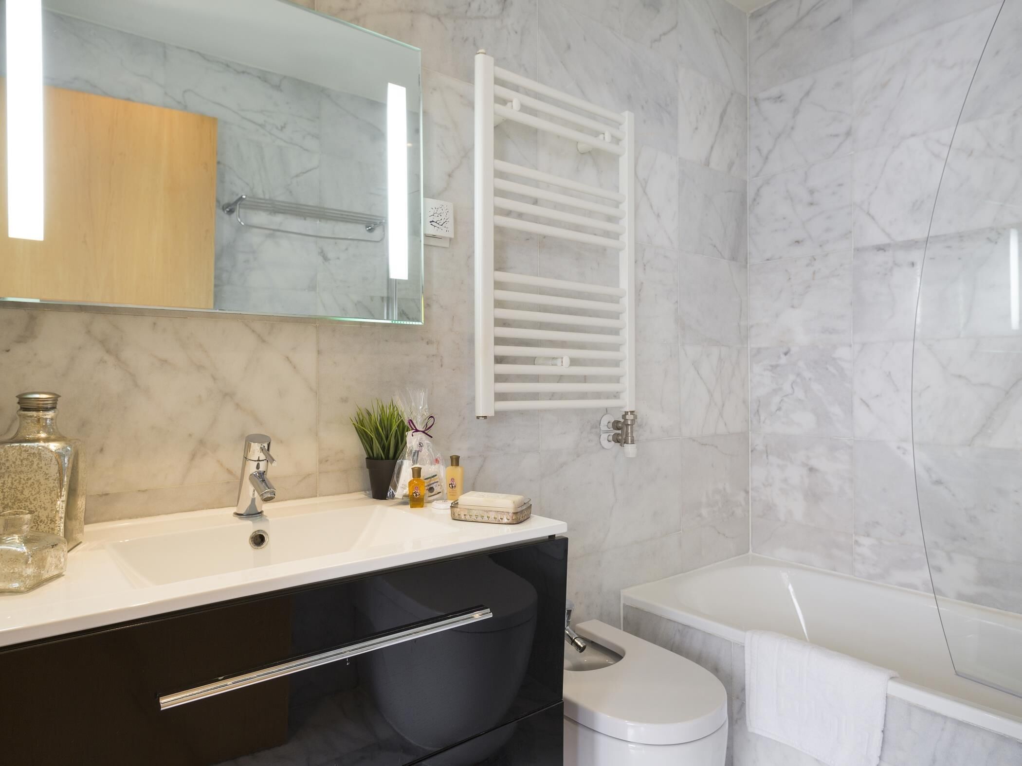 Une salle de bain moderne avec un mur en marbre, une baignoire blanche, un meuble-lavabo noir avec un miroir et un porte-serviettes.