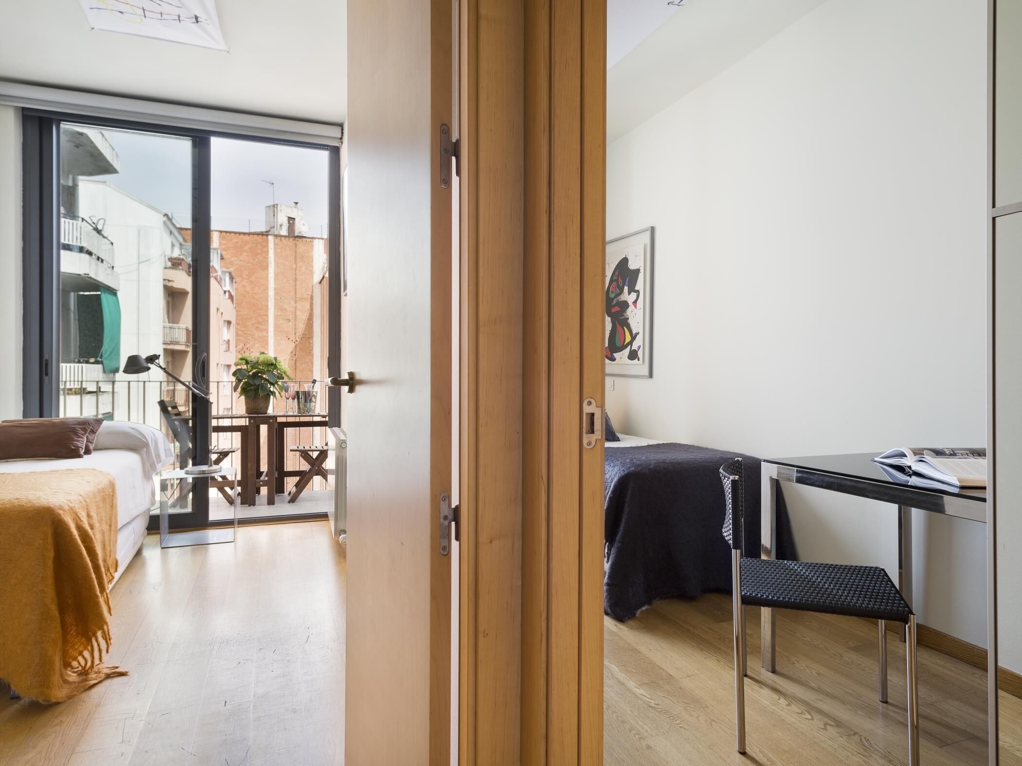A modern bedroom with a bed, a desk, and a chair. There is a balcony with a table and chairs, and a view of buildings outside.