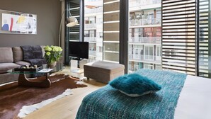 A modern living room with a sofa, a glass coffee table, a television, and a large window with blinds.