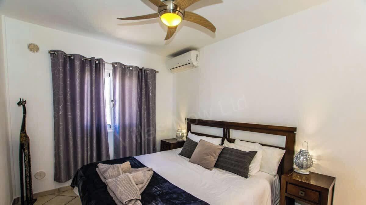 Een slaapkamer met een bed, gordijnen, een plafondventilator, een airconditioning en een nachtkastje met een lamp, plus een decoratief object aan de muur.