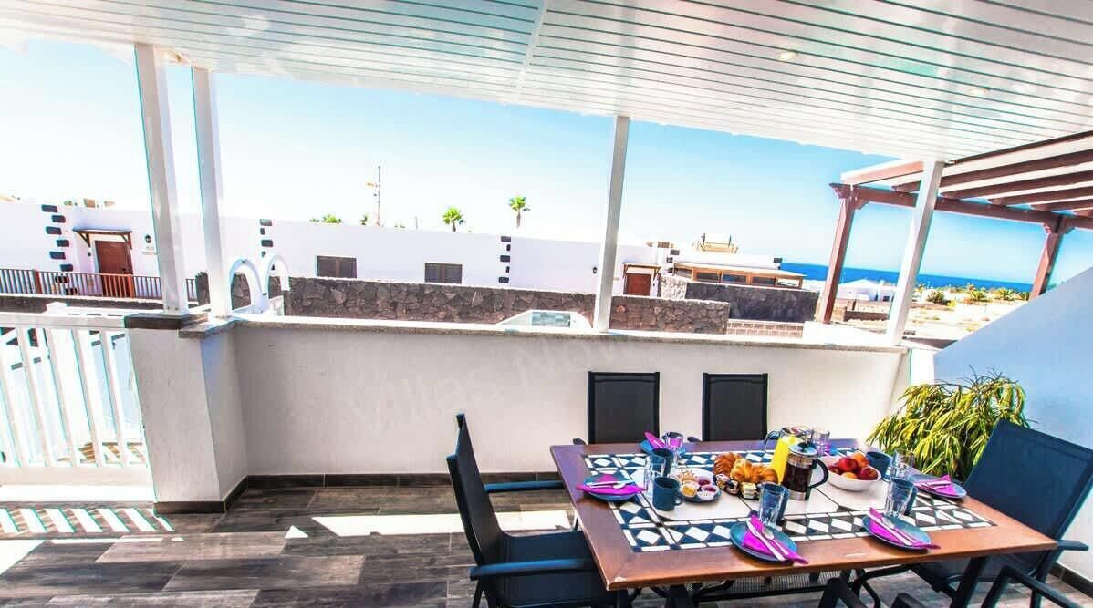 An outdoor dining area with a table set for two, a stone wall, and a view of the sea and buildings in the background.