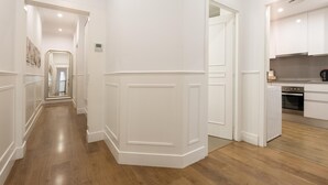 Un pasillo con paneles blancos, suelo de madera y vistas a una cocina con armarios y electrodomésticos blancos.