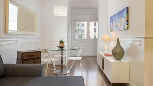Una habitación de hotel compacta con cama, una mesa de vidrio con sillas, un mueble pequeño, una lámpara y un cuadro en la pared.
