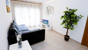 Un salon moderne avec un canapé noir, une table basse blanche, un téléviseur posé sur un meuble blanc, une plante en pot et une fenêtre ornée de rideaux légers.