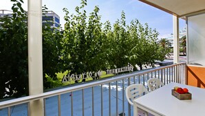 Un balcon avec une table et des chaises blanches, une rambarde en métal et une vue sur une rue bordée d’arbres.
