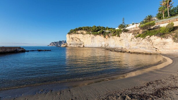 A coastal beach with a clear sky, rocky cliffs, and sparse vegetation. - Villa Bernia - PlusHolidays (Moraira)