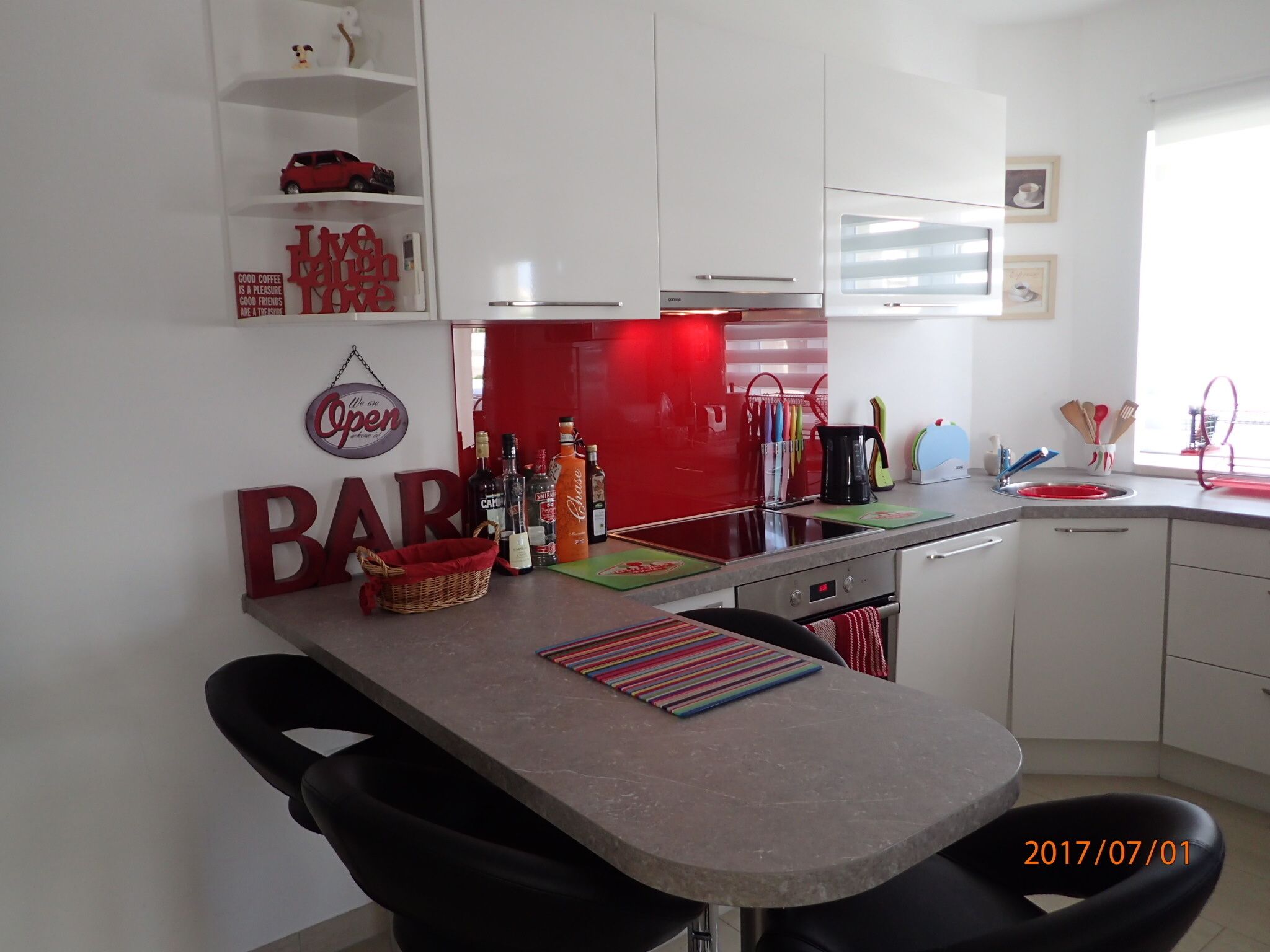 Een moderne keuken met een bar, voorzien van een aanrecht met verschillende flessen, een koffieapparaat en een rode achterwand.