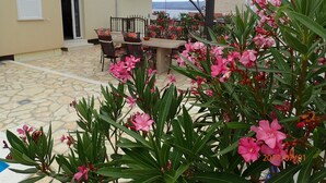 Un espace extérieur aménagé en terrasse, avec une table et des chaises, entouré de plantes en fleurs et offrant une vue sur la mer.