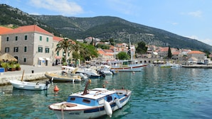 Un port de plaisance avec des bateaux amarrés, un bâtiment en pierre avec des volets verts, et une montagne en arrière-plan.