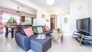 A modern living room with a grey sectional sofa, a dining table with chairs, a TV on a stand, and a kitchen in the background. - Sunset Villa - Charming Protaras Centre Getaway (Protaras)