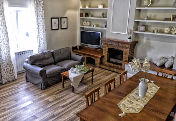 A living room with a sofa, TV, fireplace, dining table, and shelves with decorative items.